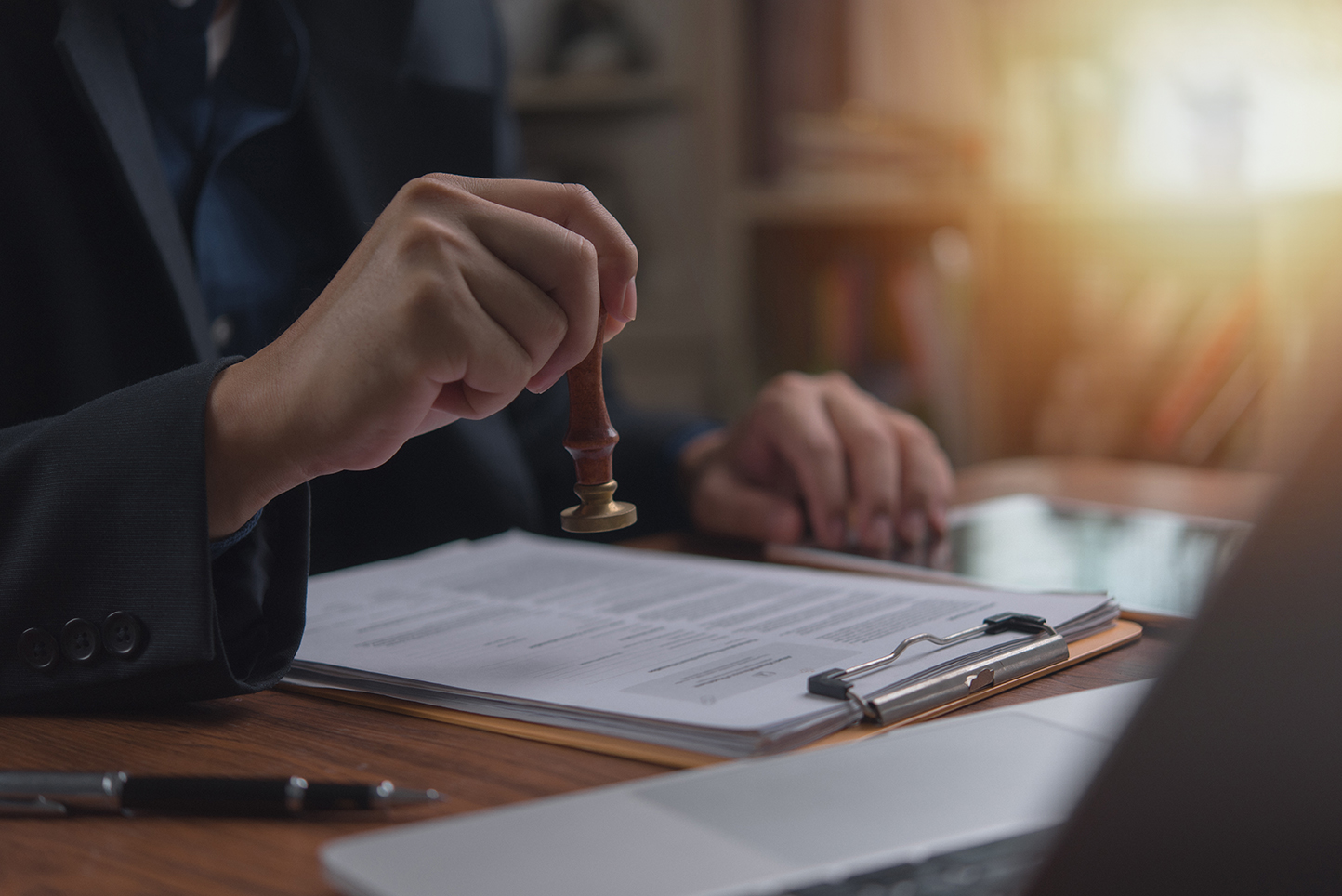 Individual in an office setting applies a stamp to a document contract legal agreement, signifying approval or verification certification permit visa next to an open laptop. permit approved photo