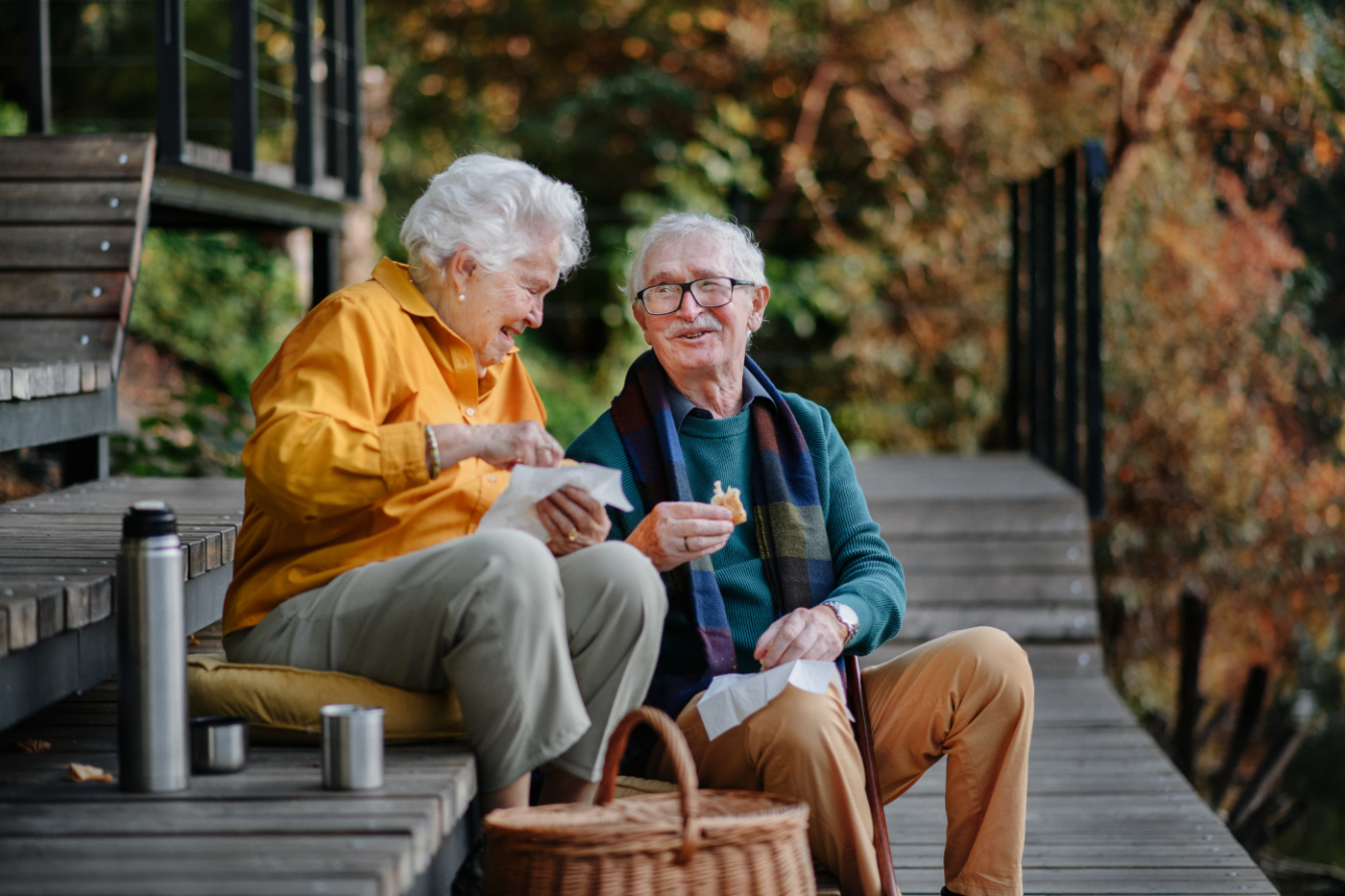 Happy senior couple in autumn clothes having picnic near lake after walk. FC IDA Senior Housing Photo